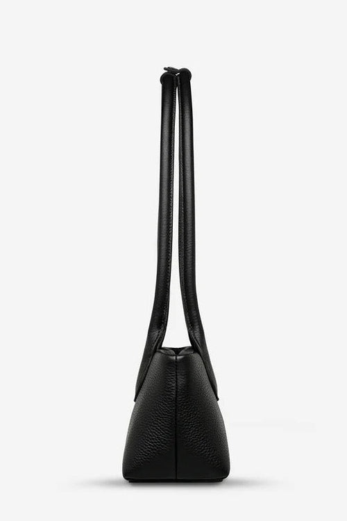 A black pebbled leather shoulder bag shown from the back, featuring a long, thin strap and a structured, tapered silhouette, set against a plain light gray background.