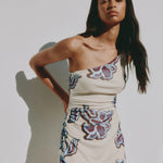 A woman modeling a one-shoulder cream-colored mini dress with a purple and blue floral pattern, standing against a light gray wall with a soft shadow.