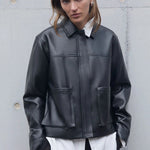 Person wearing a black leather jacket over a white shirt against a gray concrete wall.