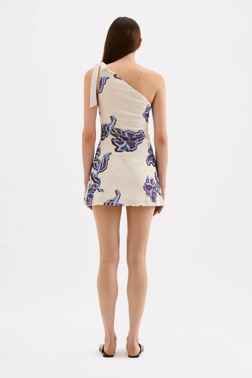 A woman viewed from the back wearing a one-shoulder mini dress with a cream base and blue and purple abstract floral print, paired with black sandals, against a plain white studio background.