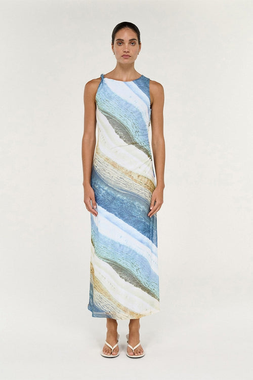 A woman modeling a sleeveless, high-neck maxi dress with an abstract, fluid wave pattern in shades of blue, beige, and white, set against a plain light background.