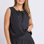 A woman modeling a sleeveless black dress with a gathered, tie-neck bodice and a flowing skirt, photographed against a plain light gray studio background.