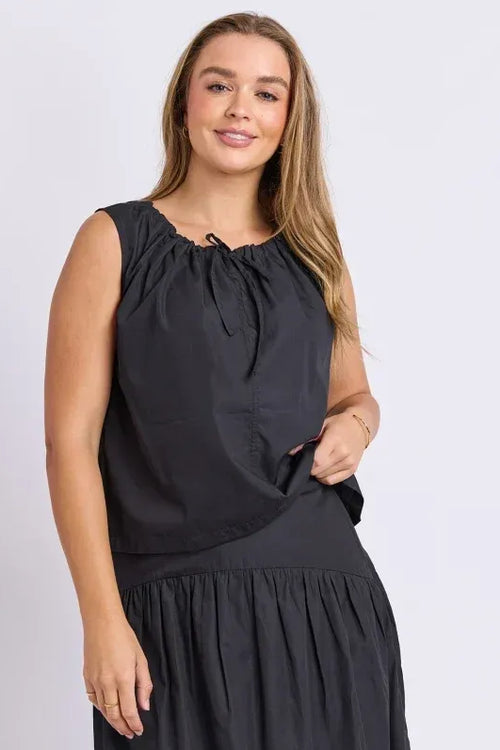 A woman modeling a sleeveless black dress with a gathered, tie-neck bodice and a flowing skirt, photographed against a plain light gray studio background.