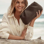 A woman with blonde hair sitting on a piece of driftwood at the beach, holding a dark brown leather pouch bag. She is wearing a cream-colored button-down shirt.