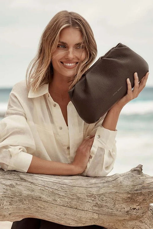 A woman with blonde hair sitting on a piece of driftwood at the beach, holding a dark brown leather pouch bag. She is wearing a cream-colored button-down shirt.