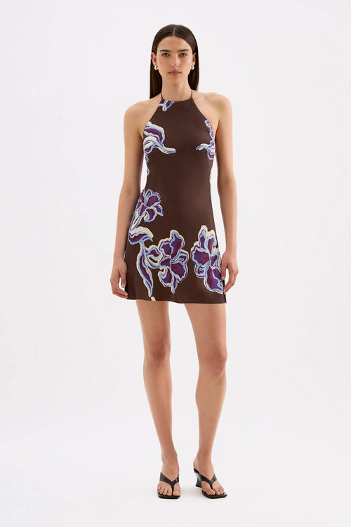 A model wearing a brown halter-neck mini dress with a purple and white floral pattern, paired with black sandals, against a plain white studio background.