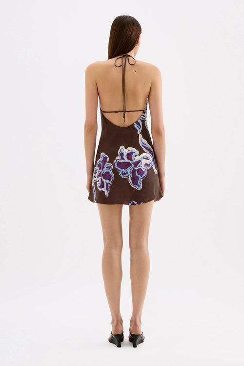 A woman viewed from the back wearing a brown halter mini dress with a purple and white floral print, paired with black heels, against a plain white studio background.