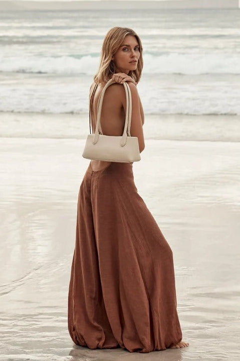 A woman stands on a beach facing away from the camera, wearing a long rust-brown maxi dress and carrying a cream-colored shoulder bag.