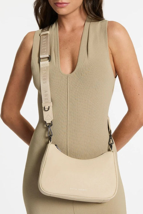 A woman wearing a beige ribbed sleeveless dress models a cream-colored Marc Jacobs crossbody bag with a matching cream strap featuring the brand name.