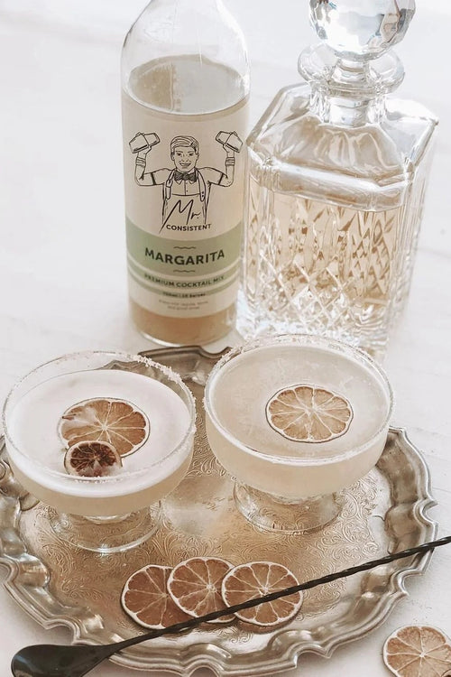 A bottle of Margarita premium cocktail mix by Mr. Consistently and a clear glass decanter sit on a silver tray beside two margarita glasses with lime wheel garnishes, on a white surface.