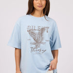 A woman modeling a light blue oversized graphic t-shirt with a distressed eagle design and the text 'SILENT Theory' on the front, paired with off-white pants against a light gray studio background.