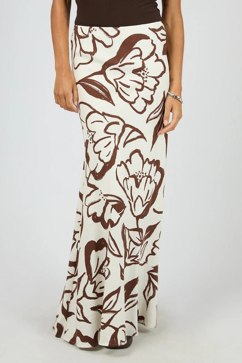 A woman wearing a long cream maxi skirt with a brown floral print, paired with a dark brown top, shown against a light gray studio background.