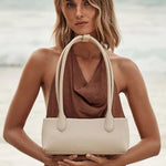 A woman standing on a beach holds a structured cream-colored leather shoulder bag with double top handles. The background shows soft-focus ocean waves and shoreline.