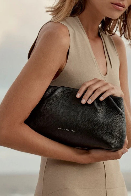 A woman in a beige sleeveless dress holds a black pebbled leather clutch bag with the brand name 'STATUS ANXIETY' printed in small white letters on the front, set against a soft, blurred outdoor background.