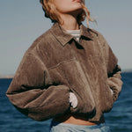 Person wearing a brown jacket and light blue pants standing by the ocean.