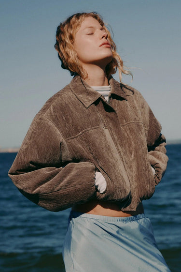 Person wearing a brown jacket and light blue pants standing by the ocean.