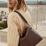 A woman wearing a yellow and white striped shirt carries a dark brown leather shoulder bag with a matching strap, set against a sunny outdoor patio background with a pool and an orange umbrella.