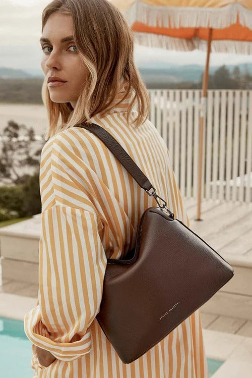 A woman wearing a yellow and white striped shirt carries a dark brown leather shoulder bag with a matching strap, set against a sunny outdoor patio background with a pool and an orange umbrella.
