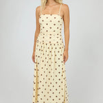 A woman modeling a cream maxi dress with brown polka dots and thin spaghetti straps, standing against a neutral light gray studio background.