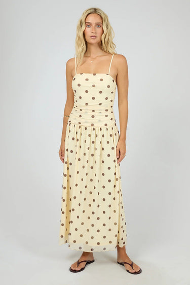 A woman modeling a cream maxi dress with brown polka dots and thin spaghetti straps, standing against a neutral light gray studio background.