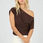 A blonde woman wearing a dark brown one-shoulder top with short sleeves and white pants, standing against a light gray studio background.