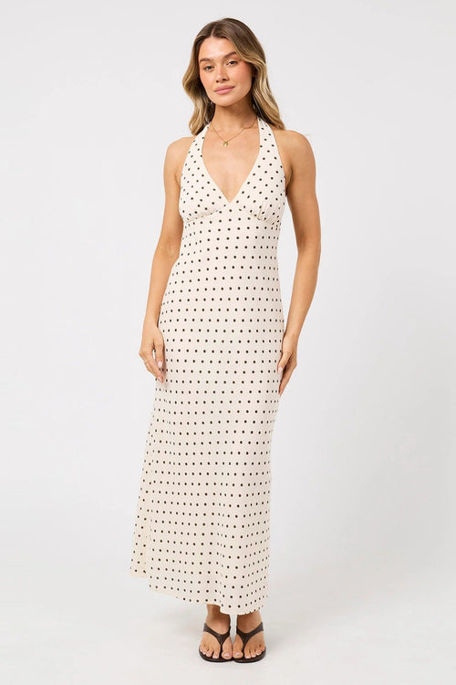 A woman modeling a sleeveless halter-neck maxi dress with a white base and small black polka dots, paired with black flat sandals against a plain light gray studio background.