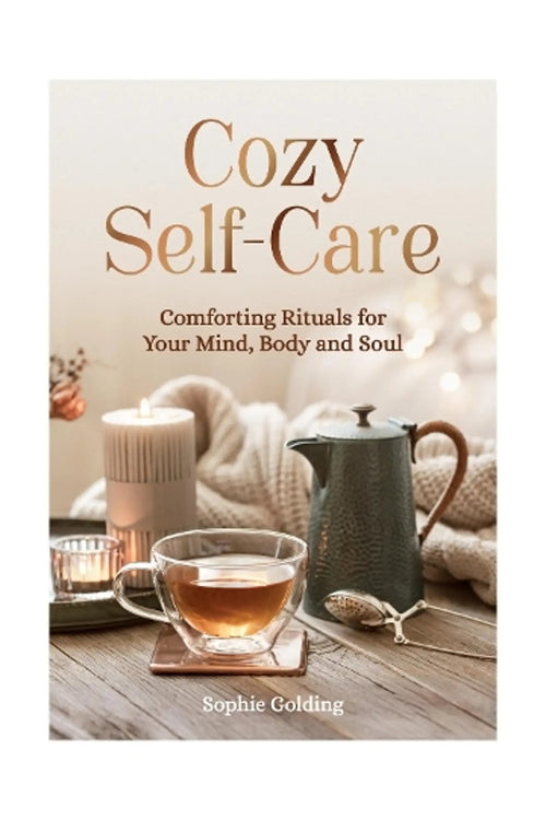 Book cover for 'Cozy Self-Care' by Sophie Golding, featuring a warm, cozy scene with a glass cup of tea, a candle, a textured metal teapot, and a knitted blanket on a wooden table.