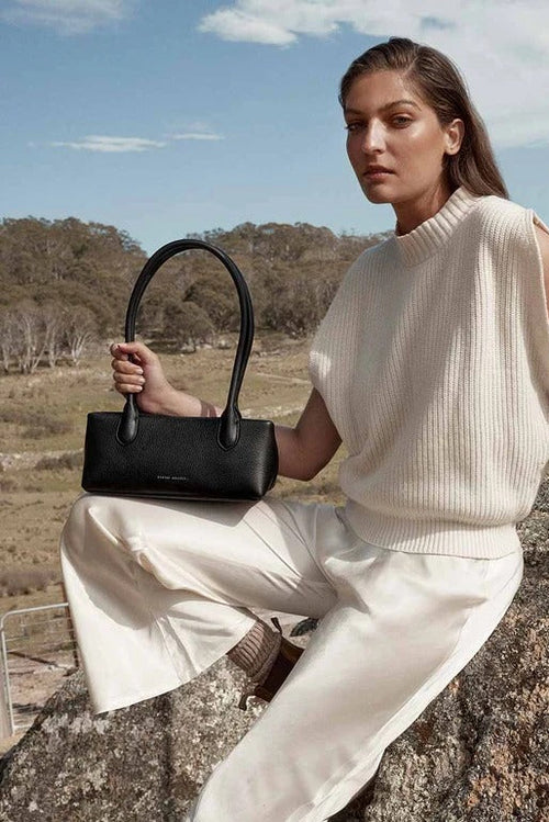Woman in a cream outfit holding a black handbag, sitting on a rock with a natural landscape in the background.