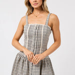 A woman modeling a sleeveless mini dress with a black and white gingham pattern, featuring a smocked bodice, thin shoulder straps, and a gathered skirt, set against a plain light gray background.