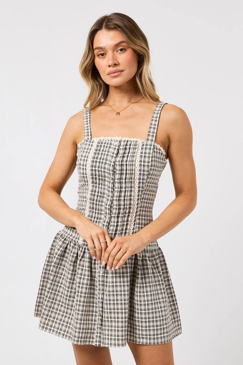 A woman modeling a sleeveless mini dress with a black and white gingham pattern, featuring a smocked bodice, thin shoulder straps, and a gathered skirt, set against a plain light gray background.