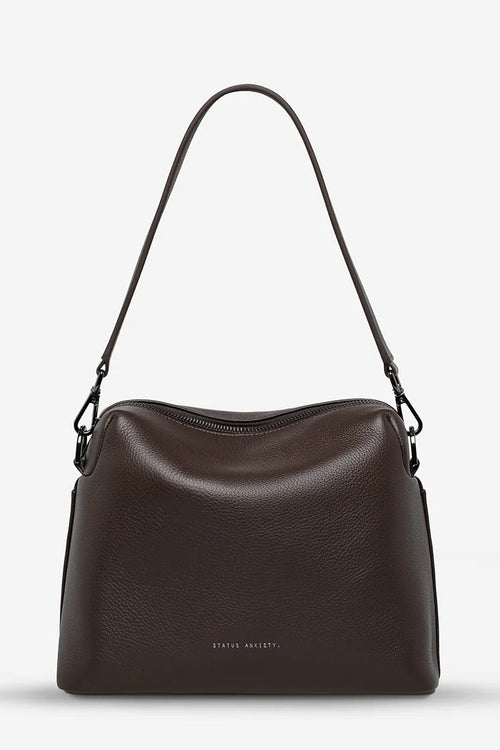 A dark brown pebbled leather shoulder bag with a single adjustable strap, shown against a plain light gray background. The brand name 'STATUS ANXIETY' is subtly printed in white near the bottom center of the bag.