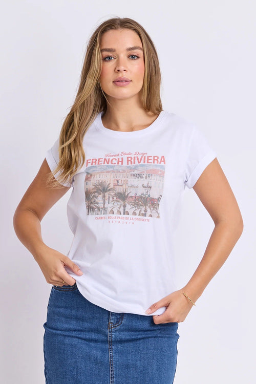 A woman modeling a white short-sleeve graphic t-shirt with a vintage-style 'French Riviera' print on the front, paired with a blue denim skirt, against a plain white studio background.