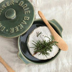 A ceramic Brie cheese warmer dish with its lid off, showing a round wheel of Brie cheese topped with a sprig of rosemary inside. A wooden spoon rests on the dish, all set on a textured cream linen background.