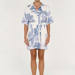 A woman modeling a short-sleeve white shirt dress with a blue floral print, featuring a button-down front, collar, and self-tie belt, set against a plain light background.