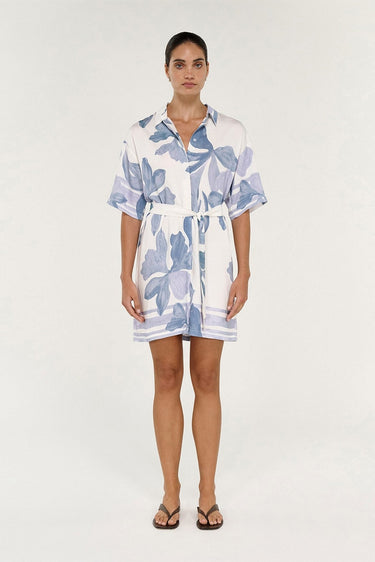 A woman modeling a short-sleeve white shirt dress with a blue floral print, featuring a button-down front, collar, and self-tie belt, set against a plain light background.