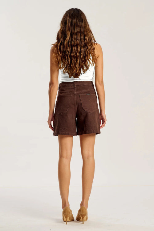 A woman viewed from behind wearing brown corduroy shorts, a white ribbed tank top, and tan high heels against a plain light gray studio background.
col_pattern