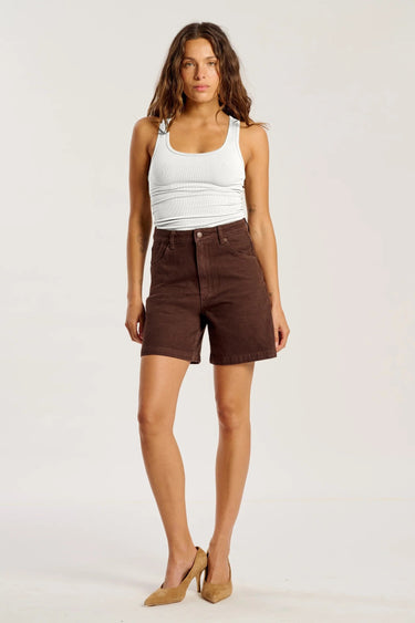A woman modeling a pair of dark brown high-waisted shorts with a white ribbed tank top and beige heels, standing against a plain white studio background.