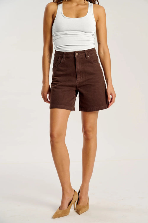 A model wearing brown high-waisted denim shorts, a white tank top, and tan pointed-toe heels, standing against a plain white studio background.