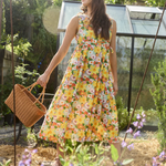 A woman stands in a sunlit garden wearing a sleeveless yellow floral dress and holding a woven basket. A greenhouse is visible in the background among green plants.
