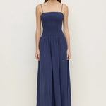 A woman modeling a navy blue maxi dress with thin spaghetti straps, a smocked bodice, and a flowing skirt, paired with black sandals against a plain white studio background.