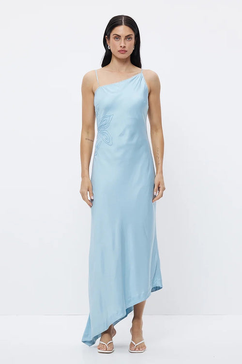 A woman modeling a light blue satin-like one-shoulder maxi dress with a butterfly embellishment on the bodice, standing against a plain white studio background.