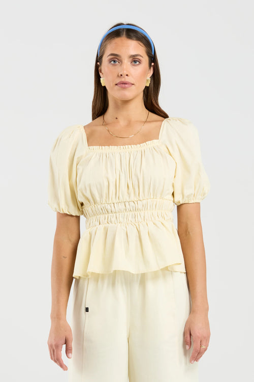 A woman modeling a pale cream puff-sleeve top with a square neckline and smocked waist, paired with matching cream pants, against a light gray studio background.