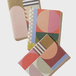 Three notebooks with abstract geometric patterns in muted pastel and earthy tones, arranged diagonally on a light gray background.