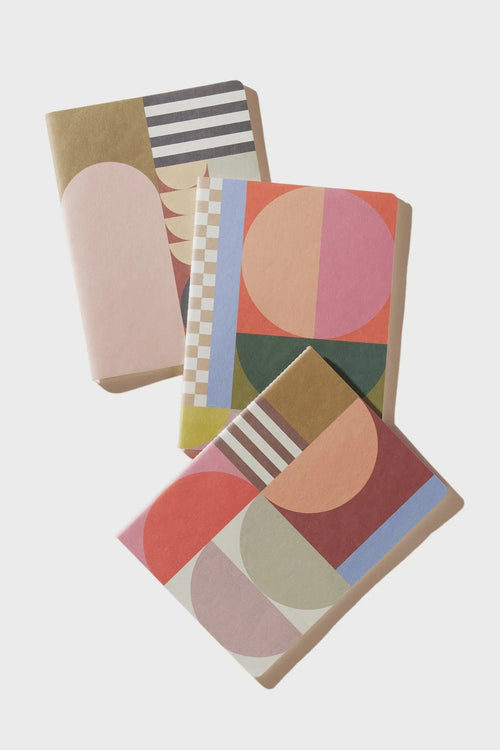 Three notebooks with abstract geometric patterns in muted pastel and earthy tones, arranged diagonally on a light gray background.