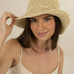 A woman wearing a beige woven straw sun hat and a cream-colored spaghetti-strap top, holding the brim of the hat against a neutral beige background.