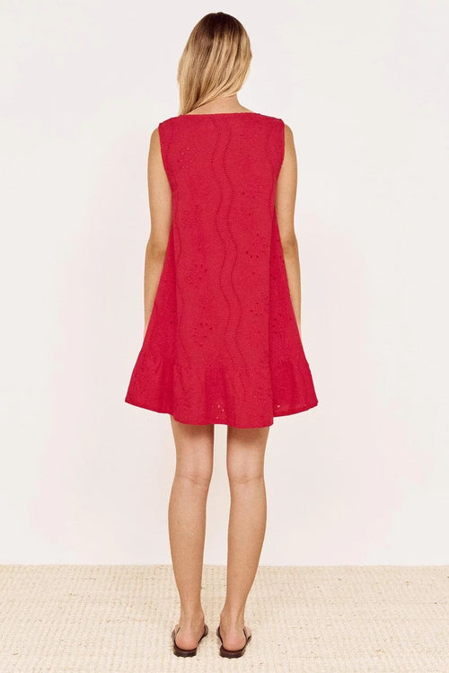 A woman viewed from behind wearing a sleeveless red eyelet dress with a flared skirt, standing against a plain off-white wall on a light woven floor.