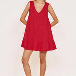 A woman modeling a sleeveless red mini dress with a V-neckline and ruffled hem, paired with dark flip-flop sandals, set against a plain off-white wall and light woven floor.