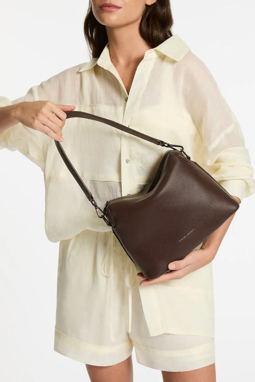 A woman wearing a cream-colored sheer shirt and shorts holds a dark brown leather shoulder bag against a plain white background.
