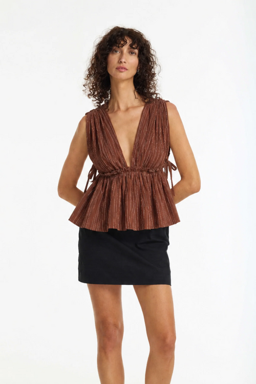 A woman modeling a sleeveless brown pleated top with a deep V-neck and gathered waist, paired with a black mini skirt, against a plain white studio background.