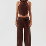 A model stands against a light gray studio background wearing a dark brown two-piece outfit consisting of a sleeveless halter-style crop top and matching wide-leg pants with a drawstring waist and metal ring detail.